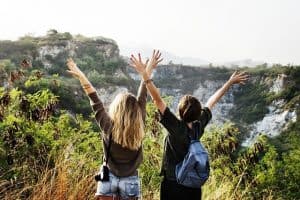 A pair of ladies with their backs to the camera w/ arms raised & outstretched while facing a mountainous view.